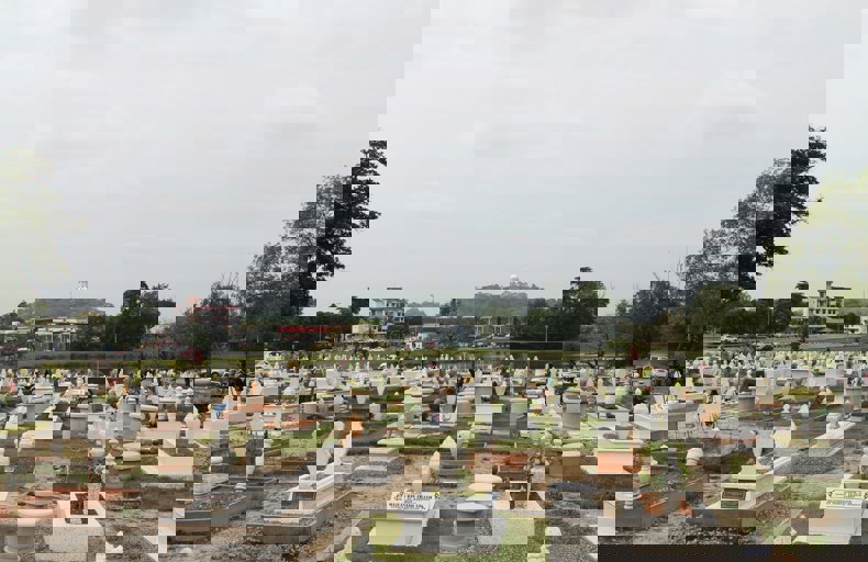 Best Sadaqah Jariyah For Deceased A wide view of a Muslim cemetery with neatly arranged graves, most of them white with decorative headstones. In the background, there are trees, buildings, and a hill with a prominent white structure on top.